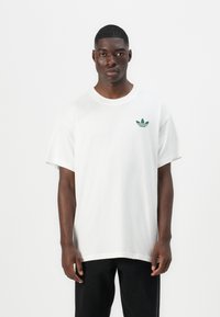 Tricou din bumbac alb cu mâneci scurte, decolteu rotund și un logo verde pe piept. Purtați-l cu pantaloni negri. Design simplu, cu o croială relaxată.