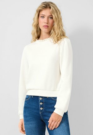 Blonde Frau mit welligem Haar, trägt ein weißes Langarmshirt mit Rüschenkragen und eine hochtaillierte blaue Jeans mit vier Knöpfen.