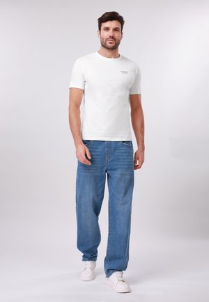 Mann som står og har på seg en hvit t-skjorte med korte ermer og tekst på brystet, løstsittende blå jeans og hvite joggesko på en ensfarget bakgrunn.