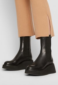 Stivaletti alla caviglia in pelle nera con suola platform spessa, caratterizzati da pannelli elastici laterali e una trama liscia, abbinati a pantaloni beige a coste.