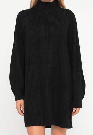 Femme portant une robe-pull noire en tricot à manches longues avec un col montant côtelé et des poignets côtelés sur fond blanc.