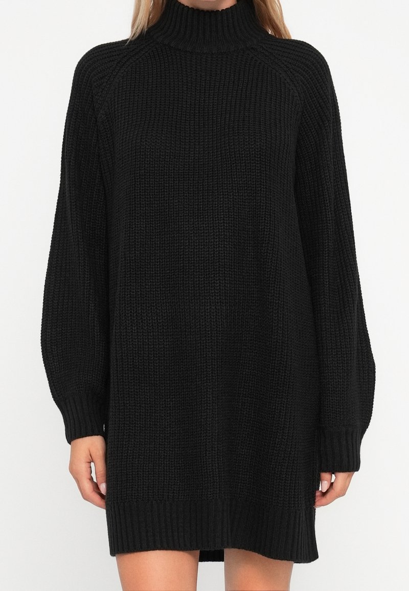 Femme portant une robe-pull noire en tricot à manches longues avec un col montant côtelé et des poignets côtelés sur fond blanc.
