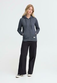 Mørkegrå zip-up hættetrøje med snøre, fleecetextur, frontlommer, båret over sorte vidde bukser og sneakers. Simpelt, afslappet design.