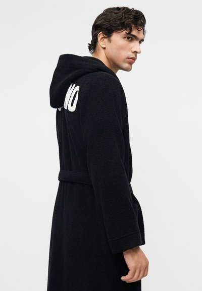 Bărbat purtând o robă neagră cu glugă, cu text alb parțial vizibil pe spate, stând pe un fundal simplu și deschis la culoare.