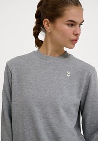 Grijze sweatshirt met een ronde hals en lange mouwen, voorzien van een gestructureerde stof en een klein chevronlogo geborduurd op de borst.