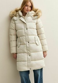 Mujer con un abrigo de plumas largo color beige con capucha de piel sintética, abrochado y con botones, de pie contra una pared clara, usando jeans azules.