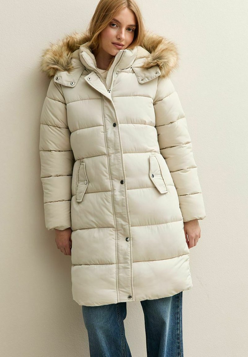 Mujer con un abrigo de plumas largo color beige con capucha de piel sintética, abrochado y con botones, de pie contra una pared clara, usando jeans azules.