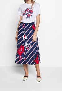 Camiseta blanca con estampado floral, combinada con una falda a rayas en azul marino con grandes diseños florales rojos. Usada con zapatos planos de color crema.