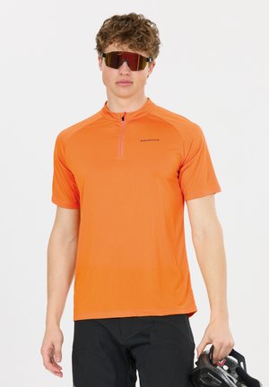 Junger Mann trägt ein orangefarbenes, kurzärmeliges Fahrradtrikot und Sonnenbrille, hält einen schwarzen Fahrradhelm in seiner rechten Hand.