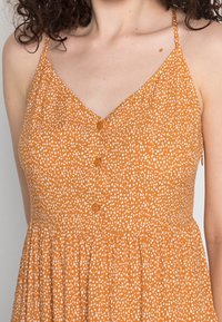 Robe orange à pois blancs, col en V, fines bretelles et détail de boutons sur le devant. Bas évasé avec texture froncée.