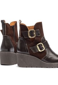 Botas tobilleras marrones con un acabado de cuero texturizado y gamuza, amplios tacones cuadrados, doble hebilla y una cremallera lateral para mayor comodidad al ponérselas.