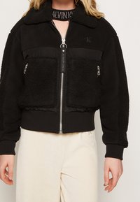 Czarna polarowa kurtka z zamkiem błyskawicznym, przednimi kieszeniami i dzianym kołnierzem. Charakteryzuje się połączeniem teksturowanej tkaniny i gładkich paneli.