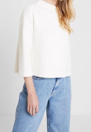 Kobieta ubrana w biały miękki sweter z rękawami do łokcia i jasnoniebieskie, wysokowcześniej dopasowane dżinsy, stojąca na tle jednolitego tła.