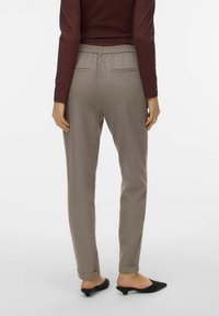 Donna che indossa pantaloni slim-fit color taupe con vita elastica e risvolti, abbinati a scarpe nere con tacco basso e senza lacci, in piedi su sfondo bianco.