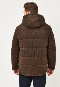 Braune gepolsterte Jacke mit weicher Textur, ausgestattet mit einer großen Kapuze und vertikaler Steppung. Der Träger hat den Rücken zur Kamera gedreht.