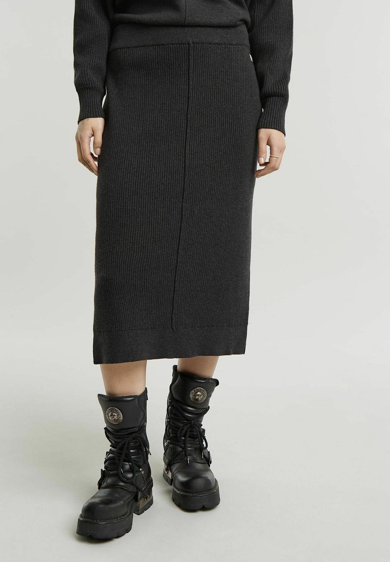 Personne portant une jupe midi côtelée en maille foncée et des bottines de combat noires à lacets avec des détails d'emblèmes métalliques, debout sur un fond clair.