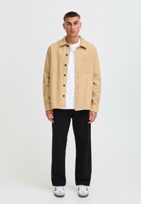 Beige corduroy shirt jacket with a buttoned front, pointed collar, single chest pocket, and black buttons, paired with black pants and sneakers.
