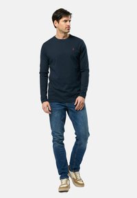 T-shirt à manches longues bleu marine en coton, arborant un petit logo rouge. Assorti à un jean bleu clair et des baskets beiges avec des accents blancs.