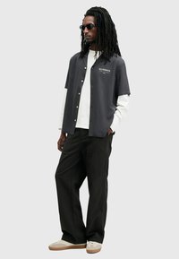 Korte mouw grijs overhemd met gouden knopen, gedragen over een witte lange mouw shirt, gecombineerd met zwarte broeken en beige sneakers.