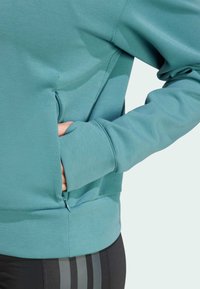 Teal sweatshirt with long sleeves, ribbed cuffs, and a side zip pocket, paired with black leggings featuring three vertical stripes.