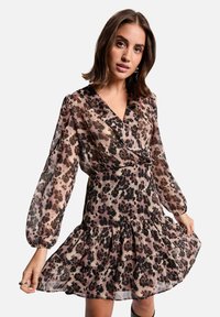 Femme portant une robe à manches longues, décolletée en V, imprimé léopard avec un ourlet évasé, tenant les bords de la jupe, se tenant devant un fond clair.