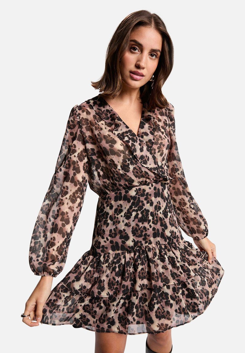 Femme portant une robe à manches longues, décolletée en V, imprimé léopard avec un ourlet évasé, tenant les bords de la jupe, se tenant devant un fond clair.