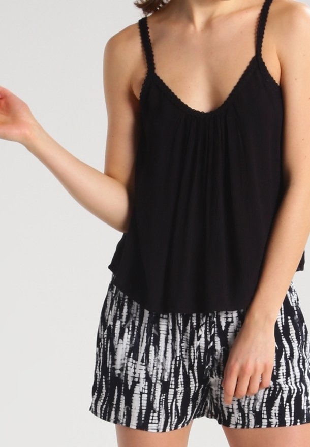 Mujer con una blusa negra sin mangas de tirantes finos y pantalones cortos con un patrón en blanco y negro, de pie contra un fondo claro y liso.