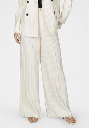 Frau trägt cremefarbene, weit geschnittene Hose mit dünnen, vertikalen Streifen, kombiniert mit passendem Blazer und weißen Sandaletten mit offenem Zehenbereich.