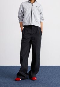 Jasnoszara kurtka zapinana na zamek z elastycznymi mankietami, zestawiona z czarnymi spodniami o szerokich nogawkach. Na stopach widoczne skarpetki w czerwono-białe wzory.