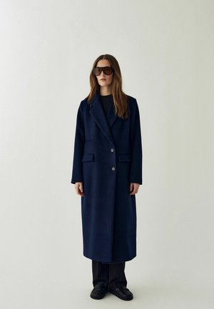 Vrouw draagt een lange donkerblauwe jas, zwarte broek, zwarte schoenen en oversized donkere zonnebril, staand tegen een effen lichte achtergrond.