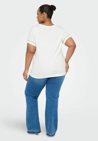 Camiseta crema de manga corta con detalles de encaje en las mangas, combinada con jeans azul de pierna acampanada con un dobladillo deshilachado, vista desde la parte posterior.