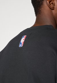 T-shirt de algodão preto com decote redondo. Na parte de trás, exibe um patch colorido do logotipo da NBA no centro superior. Textura suave.