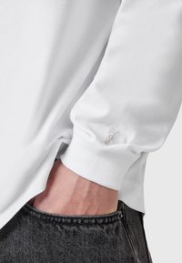 Weißes Langarmhemd mit gerippten Bündchen, versehen mit einem dezenten gestickten Logo in der Nähe des Handgelenks. Weicher Stoff und lässige Passform. Kombiniert mit schwarzen Jeans.