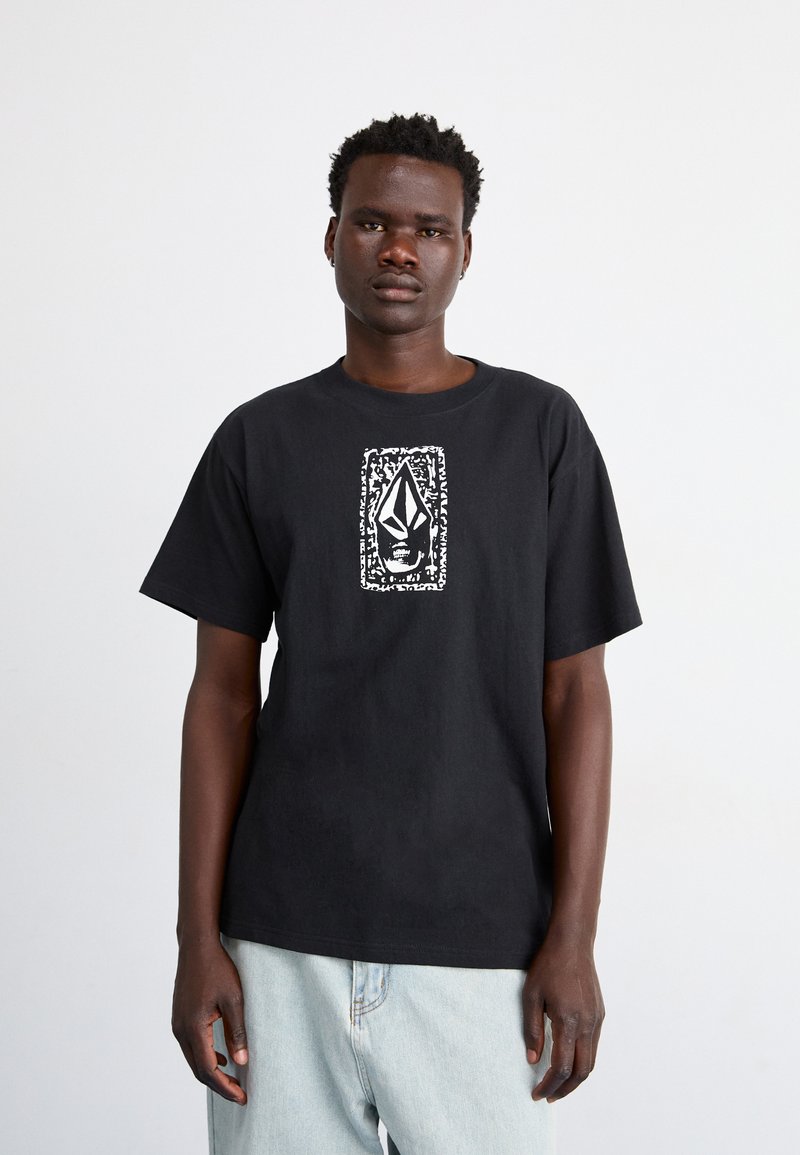 Camiseta de algodón negra con un gráfico central blanco de diseño estilizado, mangas cortas y un corte relajado. Lleva puestos unos vaqueros de un azul claro.