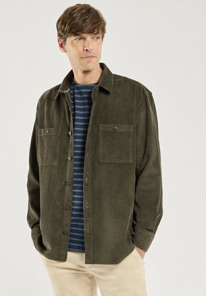 Homme portant une chemise en velours côtelé vert olive déboutonnée par-dessus un t-shirt rayé bleu marine avec un pantalon beige, mains dans les poches, regardant vers la gauche.