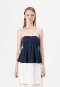 Navy strapless top with a fitted bodice and flared peplum hem, featuring ribbed texture. Model paired it with a white embroidered skirt.