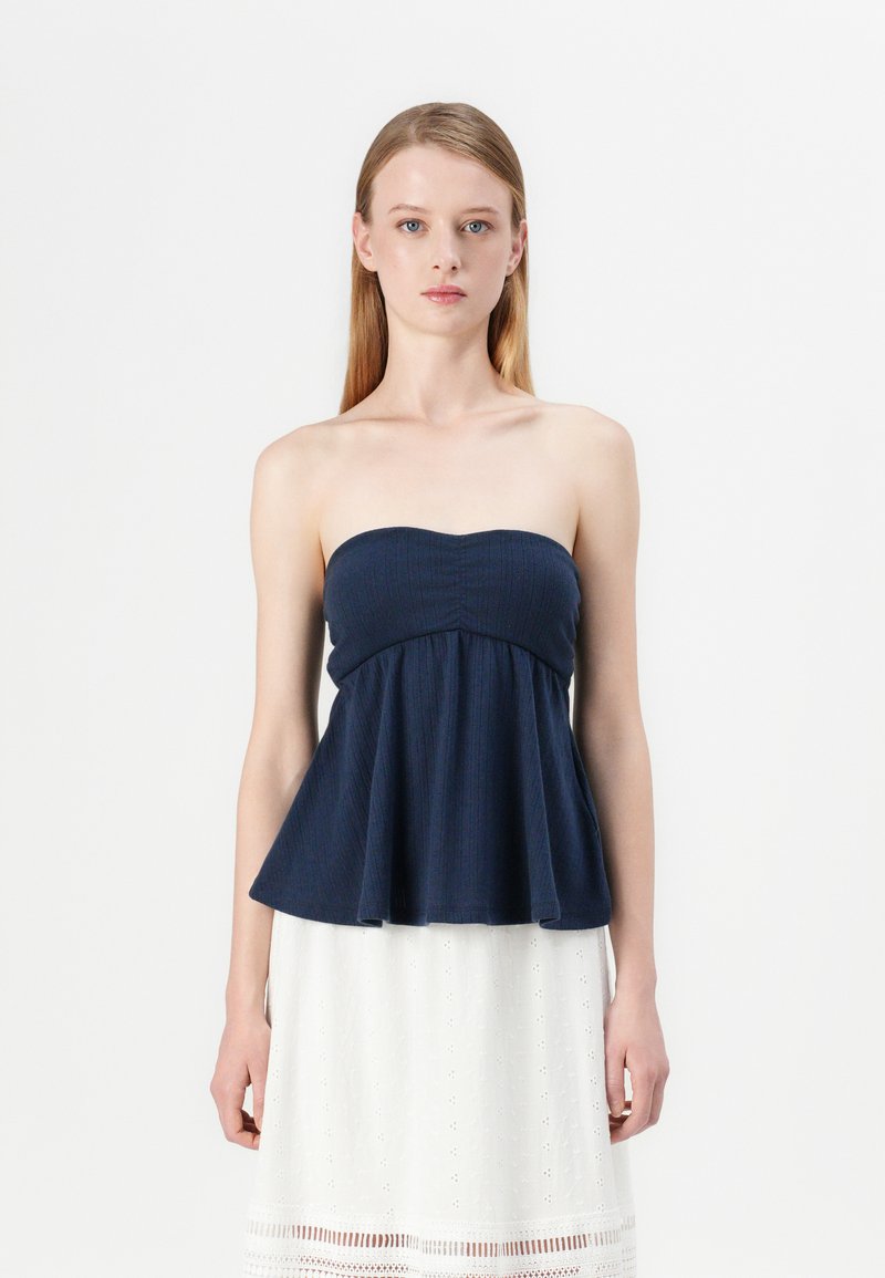 Navy strapless top with a fitted bodice and flared peplum hem, featuring ribbed texture. Model paired it with a white embroidered skirt.