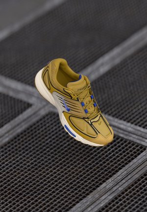 Chaussure de running Nike jaune moutarde avec des accents bleus et une semelle blanche, positionnée au-dessus d’un fond en grille métallique.