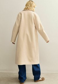 Persona con cabello rubio hasta los hombros, vistiendo un abrigo largo beige, jeans de mezclilla azul oscuro y zapatillas marrones, de pie frente a una pared lisa.