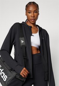 Black athletic zip-up hoodie with long sleeves, paired with a white sports bra and black high-waisted leggings, holding a black adidas gym bag.