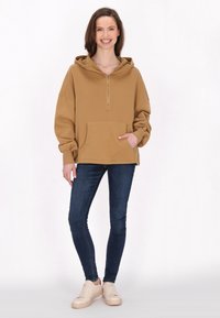 Brauner Kapuzenpullover mit halbem Reißverschluss, Fronttasche und lockerer Passform, kombiniert mit dunklen Slim-Fit-Jeans und cremefarbenen Sneakers. Mattes Material.