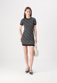 Robe en tricot noir et blanc avec un imprimé logo répété, manches courtes et silhouette ajustée. Le mannequin porte un sac crème et des talons noirs.