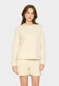 Conjunto de sudadera y pantalones cortos de color crema, hecho de una tela suave; presenta un sutil bordado de "Sonrisa" en el pecho y un diseño de corte relajado.