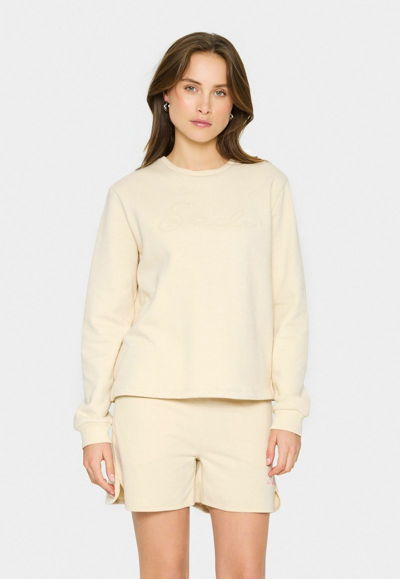 Conjunto de sudadera y pantalones cortos de color crema, hecho de una tela suave; presenta un sutil bordado de "Sonrisa" en el pecho y un diseño de corte relajado.