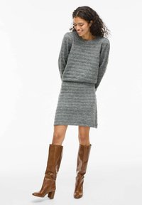 Suéter de punto gris con un patrón texturizado, combinado con una falda de punto a juego. Botas de cuero marrón hasta la rodilla con un tacón grueso.