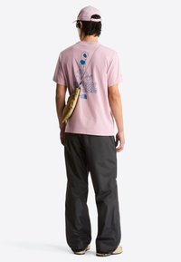 T-shirt en coton rose clair avec imprimé graphique au dos, associé à un pantalon imperméable noir. Les accessoires incluent un sac bandoulière beige et une casquette.