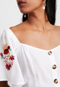 Blusa blanca, con los hombros al aire, con bordado floral en rojo, amarillo y verde. Presenta grandes botones redondos y una tela suave y texturizada.