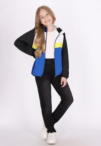 Farverig jakke i blå, sort og hvid med en gul streg; blødt stof, lynlås lukning, hætte-design. Båret med sorte jeans og hvide sko.