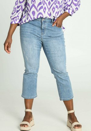 Femme portant un jean court bleu clair, un chemisier violet à motifs et des sandales compensées beige, debout avec une main dans la poche.