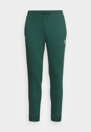 Grønne sweatpants med tætsiddende pasform, snøre i taljen og lille hvid Converse-logo på venstre lår, vist på en ensfarvet baggrund.